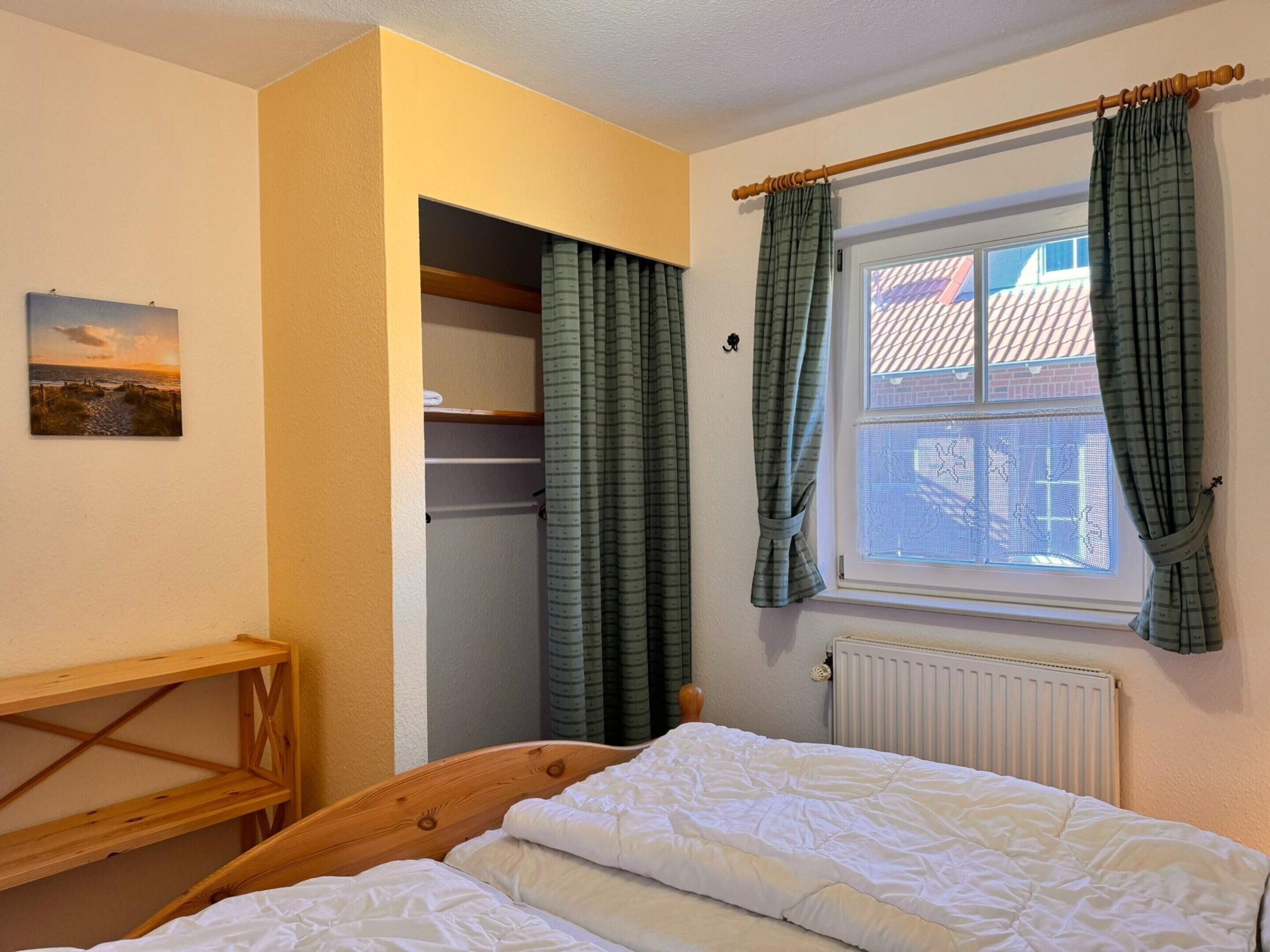 A bedroom with a wooden bed, a window with curtains, a radiator, and a shelf with a picture.