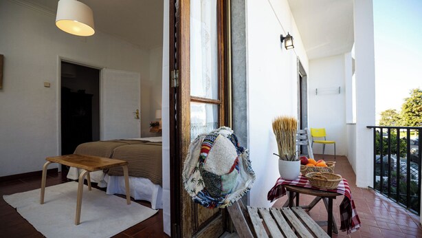 Une pièce comprenant un lit, une table en bois, un banc et un balcon avec une chaise et un panier de fruits.