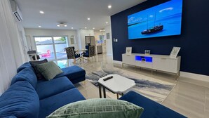 A modern living room with a blue sofa, a small white table, and a TV displaying a boat on water. - 5 Star Tropical Pool Near Beach & FLL (Pompano Beach)