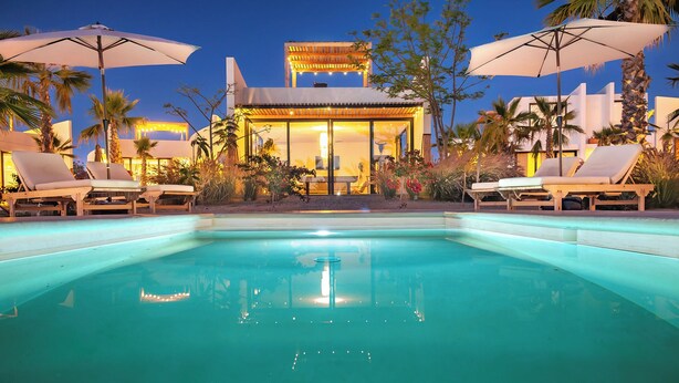 A poolside area with lounge chairs and umbrellas, a well-lit building with a pool in the background, and a clear night sky. - Casa Tove, a cosy Bungalow in El Pescadero with wifi, yoga deck and pool. (El Pescadero)