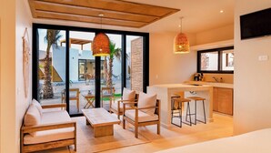 A modern living room with a sofa, coffee table, and chairs, a kitchen area with bar stools, and large windows offering a view of palm trees and a building. - Casa Tove, a cosy Bungalow in El Pescadero with wifi, yoga deck and pool. (El Pescadero)