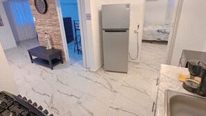 A modern kitchen with a gas stove, a refrigerator, and a sink. There is a dining area with a table and chairs, and a view of a bedroom with a bed and curtains.
