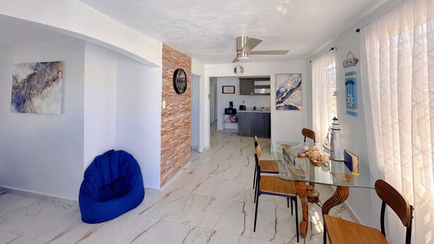 A dining area with a glass table, wooden chairs, a blue bean bag, and a kitchen in the background.