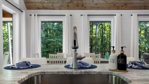 A kitchen with a stainless steel sink, white cabinetry, and large windows offering a view of a forested area.