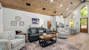 A spacious living room with a wooden ceiling, a black sofa, patterned armchairs, a coffee table, and a kitchen in the background.