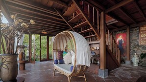 A room with a wicker swing chair, a stone wall with artwork, and a view of greenery outside. - Loid's di Lempuyang (Karangasem)