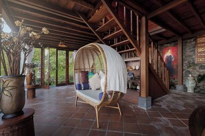 A room with a wicker swing chair, a stone wall with artwork, and a view of greenery outside. - Loid's di Lempuyang (Karangasem)