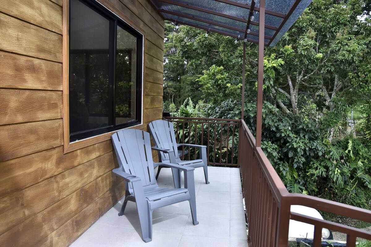 Pared exterior de madera con un ventanal grande y dos sillas grises en un balcón con barandilla de metal.