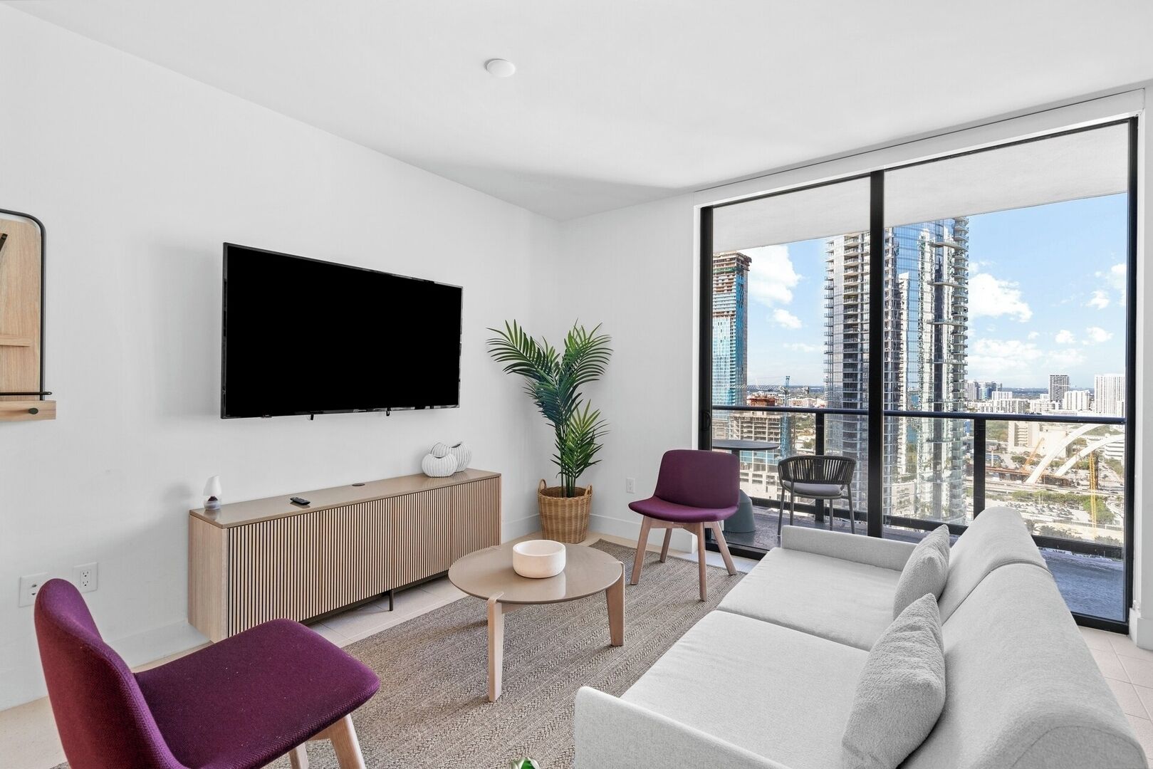A modern living room with a large TV, comfortable seating, a plant, and a view of a cityscape through large windows.