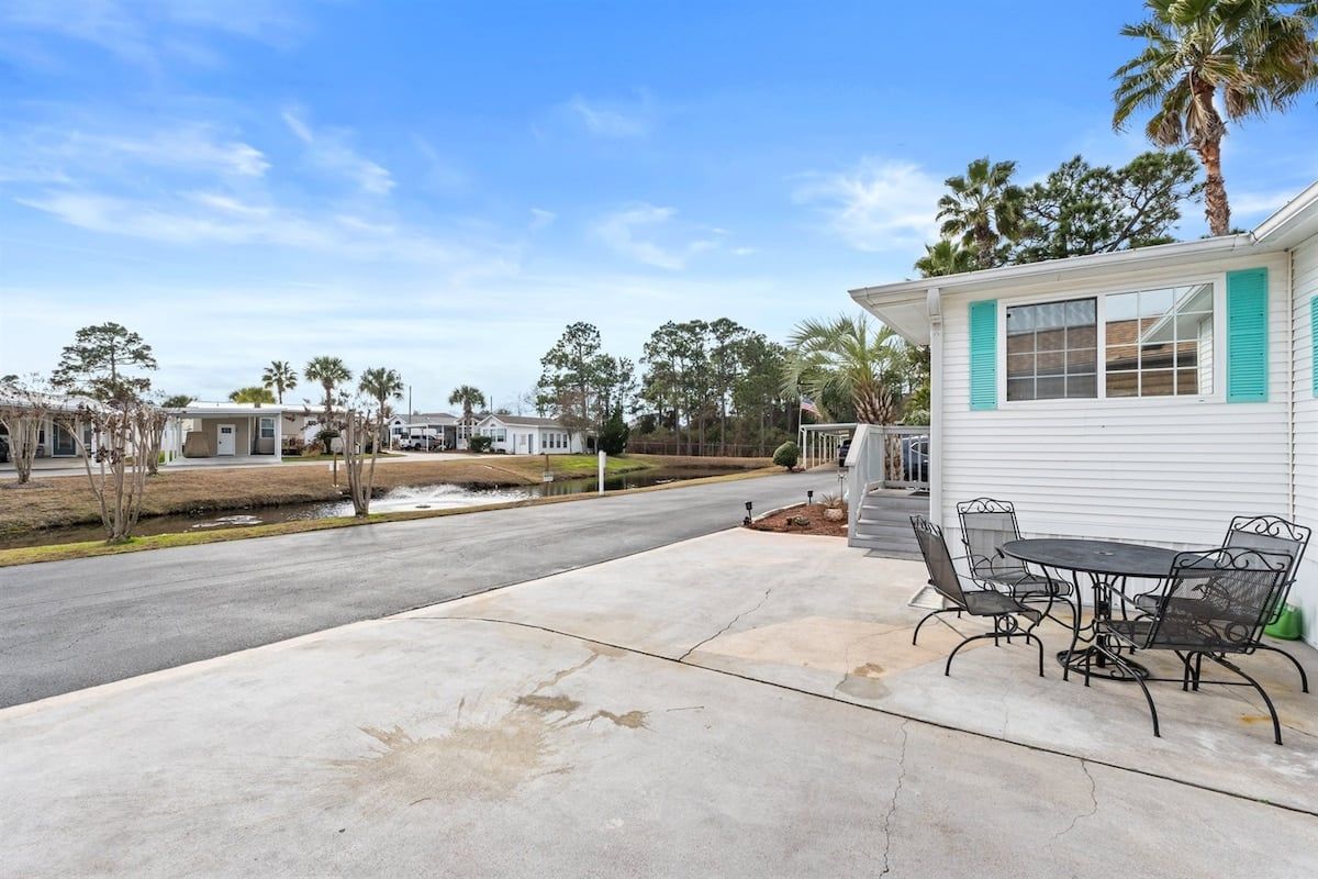 A residential property with a paved driveway, a patio with outdoor furniture, and a white building with green shutters.