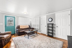 A living room with a brown leather sofa, a glass coffee table, and a patterned rug. There is a teal door, white walls, and a black bookshelf. - 3b 3ba Home, Pet allowed, 2 Pools, Hot Tub, Gym (Panama City Beach)