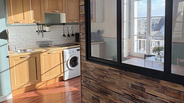 A modern kitchen with wooden cabinets, a built-in washing machine, and a large window offering a view of the outside. - Bright apartment near the quays, ideal for discovering Quimper! (Quimper)