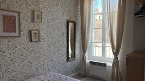 A bedroom with floral wallpaper, a bed with a patterned bedspread, a window with sheer curtains, and a wooden dresser. - Bright apartment near the quays, ideal for discovering Quimper! (Quimper)