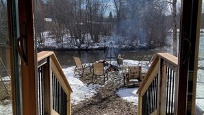 Une terrasse en bois menant à une rivière, avec des chaises et un foyer, le tout entouré d’arbres et de neige.