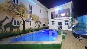 Zona de piscina exterior con un mural de animales en la pared, una cancha de tenis y un edificio de varios pisos.