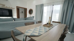 A modern, well-lit room with a long wooden dining table, a sofa set, a flat-screen TV, and a large window with curtains.