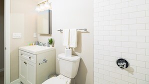 Casa de banho com lavatório branco, espelho, sanita, barra de toalhas e candeeiro fixado na parede.