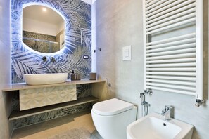 Ein modernes Badezimmer mit einem großen, ovalen Spiegel, einem weißen Waschbecken, einer Toilette und einem Bidet.
