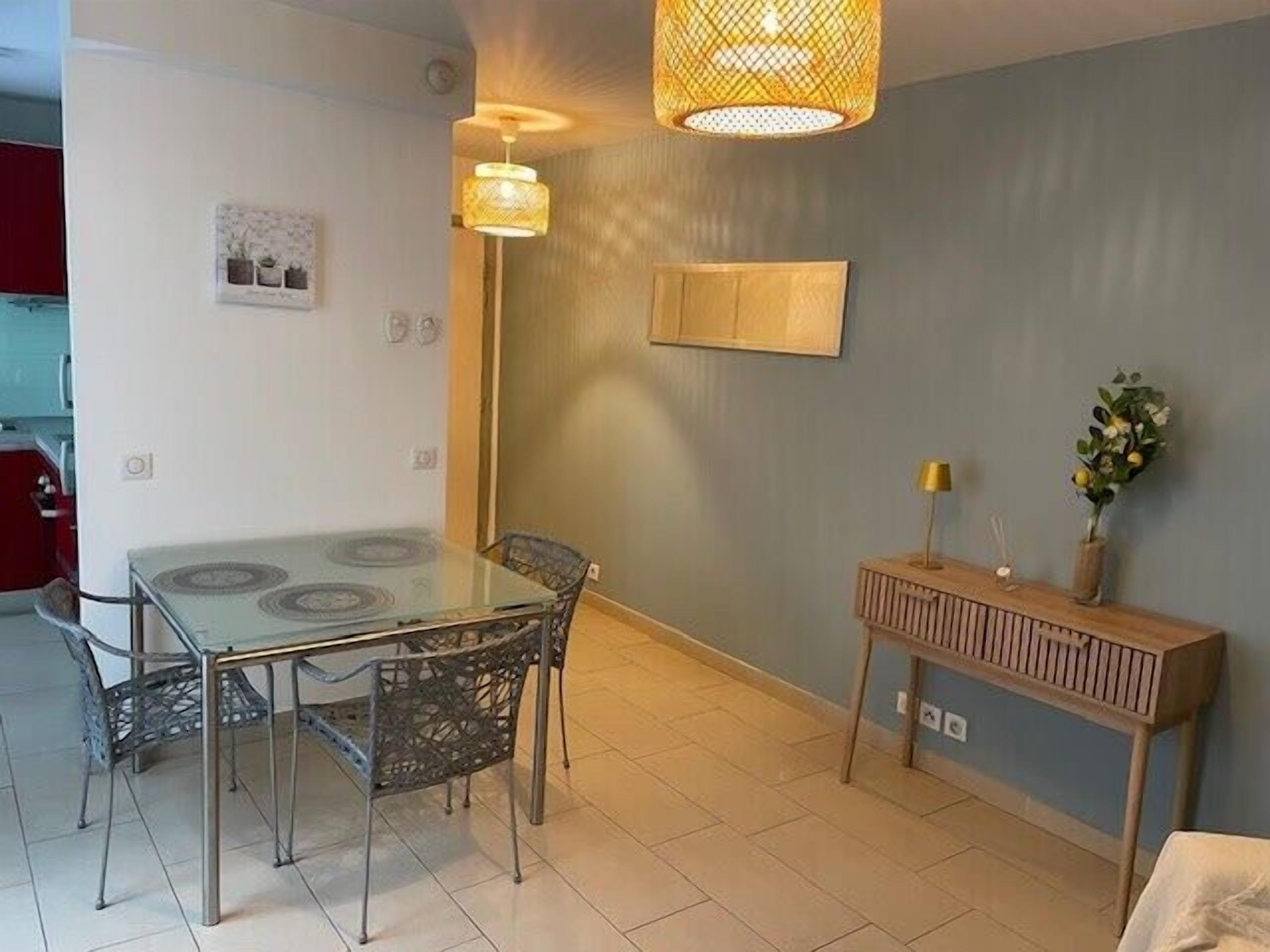A dining area with a glass table and metal chairs, a wooden sideboard with a lamp, and a wall-mounted picture.