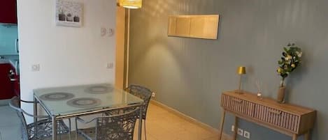 A dining area with a glass table and metal chairs, a wooden sideboard with a lamp, and a wall-mounted picture.