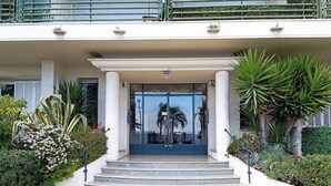 Ingresso di un hotel con porta in vetro, gradini e vegetazione circostante.