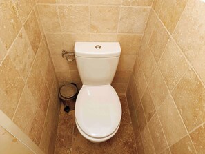 Un inodoro en un baño con baldosas color beige y un pequeño cubo de basura a su lado.
