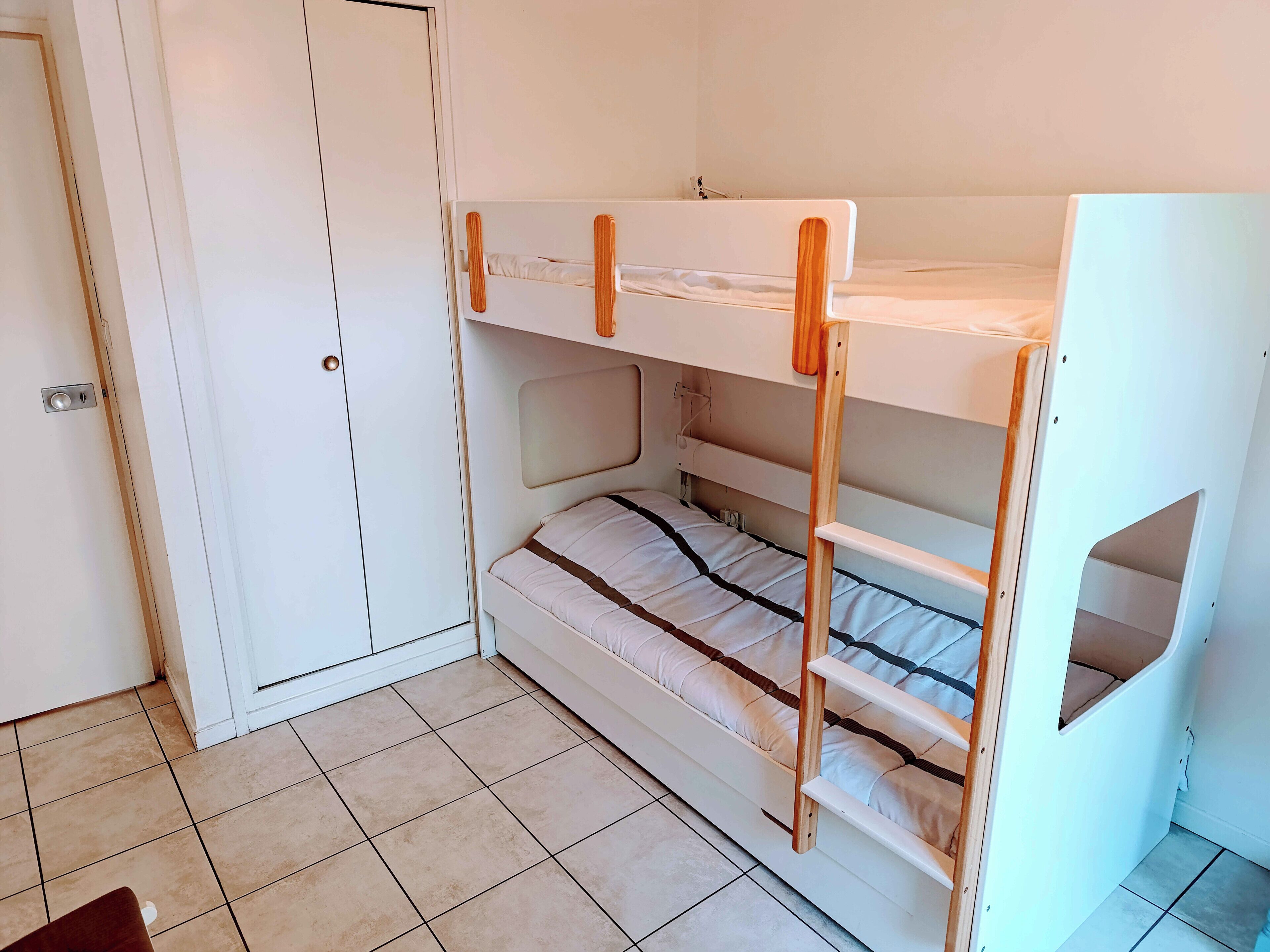 Una cama litera con estructura de madera y colchón blanco, una puerta blanca y suelo de baldosas.