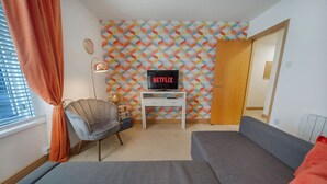 A cozy room with a grey sofa, a chair, a small white table with a TV displaying 'NETFLIX', and a colorful geometric-patterned wall. - Guild Street Apartment | Grampian Lettings Ltd (Aberdeen)