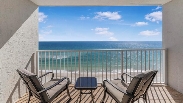 Ein Balkon mit zwei Stühlen und einem kleinen Tisch, mit Blick auf einen Strand mit klarem, blauem Wasser und einem strahlend blauen Himmel.