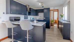 A modern kitchen with dark cabinets, a central island with bar stools, and a view of the adjacent dining area. - 1 Bed in Southcott (oc-t30813) (Okehampton Hamlets)