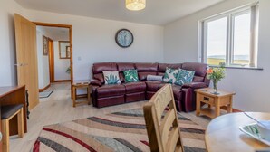 A living room with a leather sofa, a wooden coffee table, a dining table, and a large window. - 1 Bed in Southcott (oc-t30813) (Okehampton Hamlets)