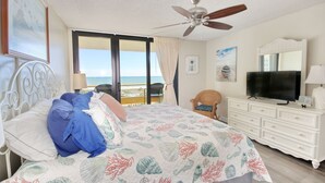 A bedroom with a bed, a television, a dresser, and a balcony with a view of the sea.
