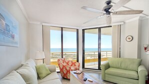 A living room with a sofa, armchair, and coffee table, a ceiling fan, and large windows with a view of the sea.
