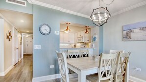 A dining area with a wooden table and white chairs, a kitchen with white cabinets and copper pendant lights, and a wall clock.