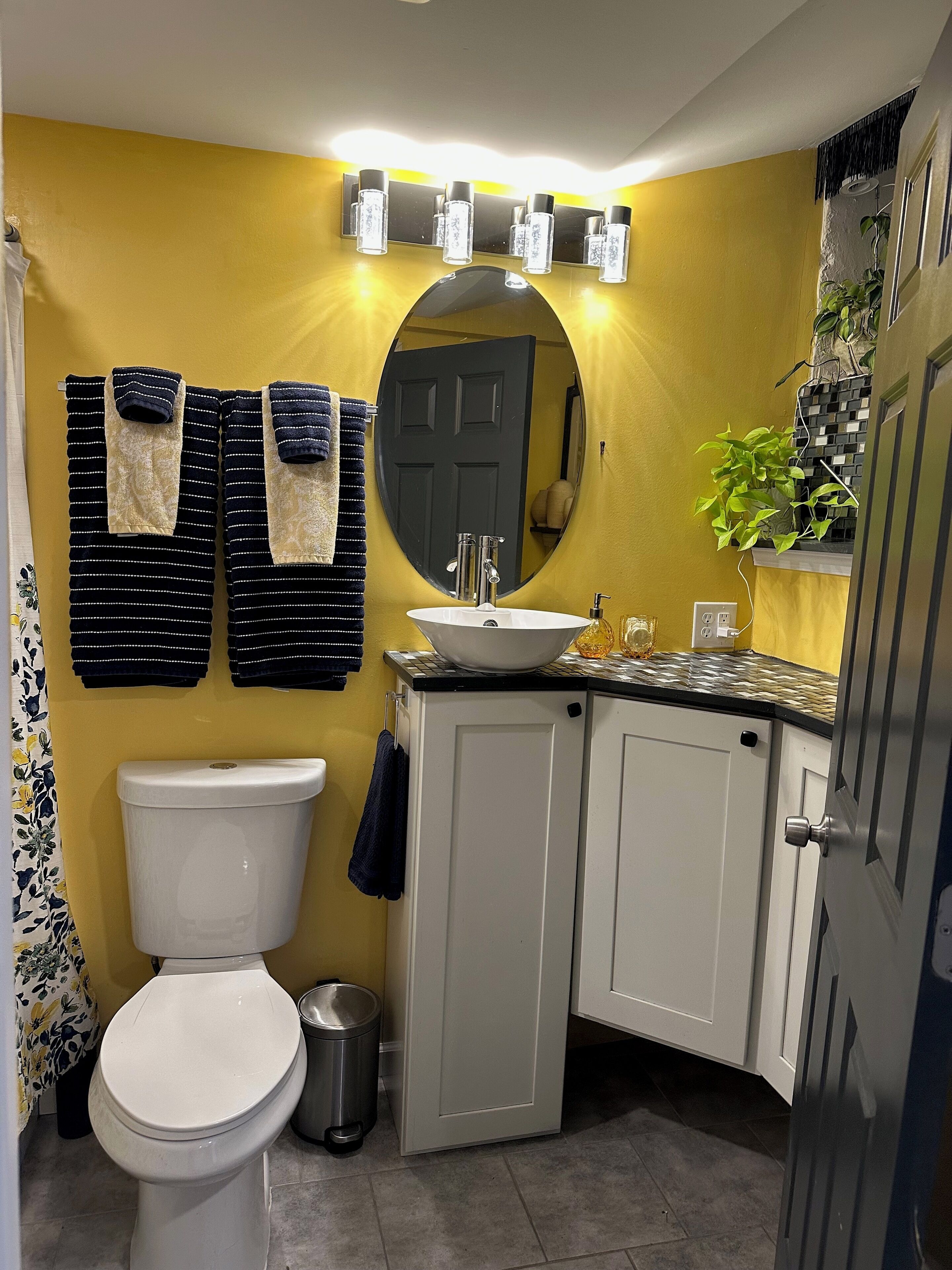 A bathroom with a yellow wall, a white toilet, a sink with a mirror above it, and a cabinet with a plant on top.