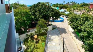 Une vue depuis un balcon donnant sur une plage bordée d’arbres, avec une piscine bleue et un ciel dégagé.