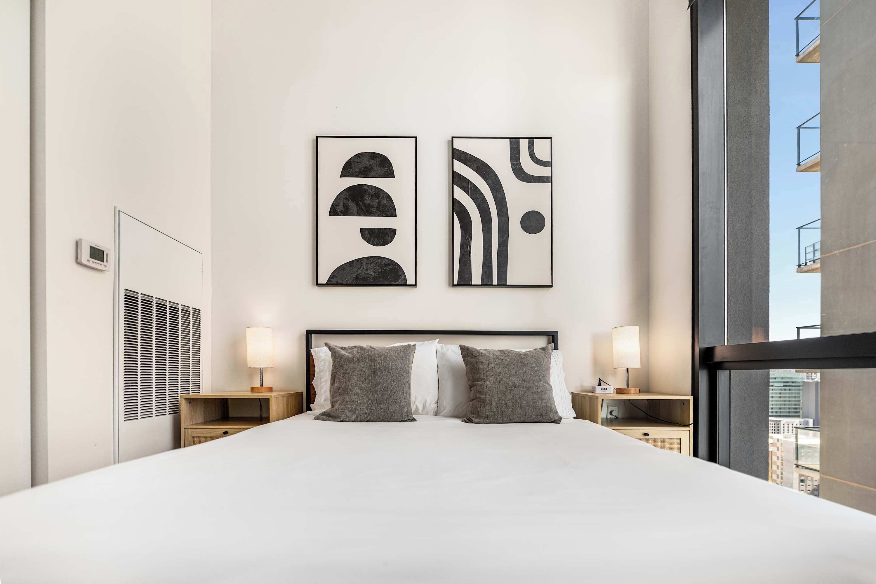 A modern hotel room with a large bed, two bedside tables with lamps, abstract wall art, and a view of the city through a large window.
