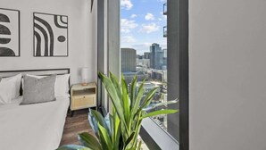 A modern bedroom with a city view, a bed with white linens, a bedside table with a lamp, and a large potted plant. - Boulevard Stays Modern Highrise STU Sky Pool, Gym 01 (Phoenix)