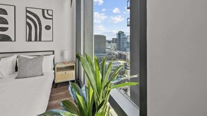 A modern bedroom with a city view, a bed with white bedding, a nightstand, and a potted plant. - Boulevard Stays Modern Highrise STU Sky Pool, Gym 05 (Phoenix)