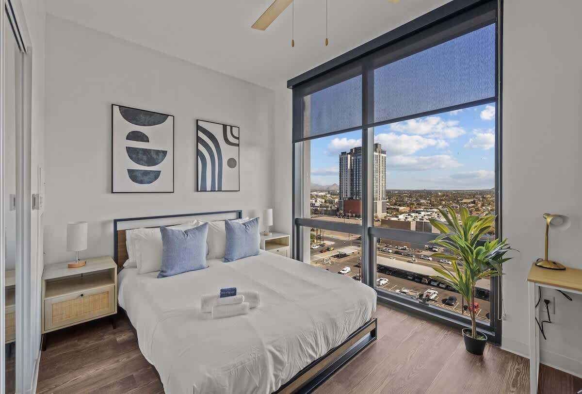 A modern bedroom with a large window, a bed with white linens, two bedside tables, and abstract wall art.