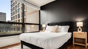 Quarto moderno com uma cama grande, duas mesas de cabeceira com luminárias, vista da cidade através de uma janela ampla e uma parede de destaque escura.