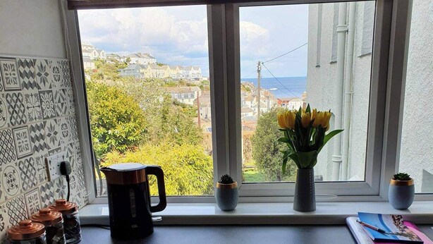 A kitchen with a window view of a coastal town, a coffee maker, a vase with flowers, and a notebook on the counter. - Seaview-petsok-patio-1br (St Austell)