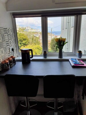 Een keuken met uitzicht op een kustplaats, een koffiezetapparaat, een vaas met bloemen en een notitieboekje op het aanrecht.