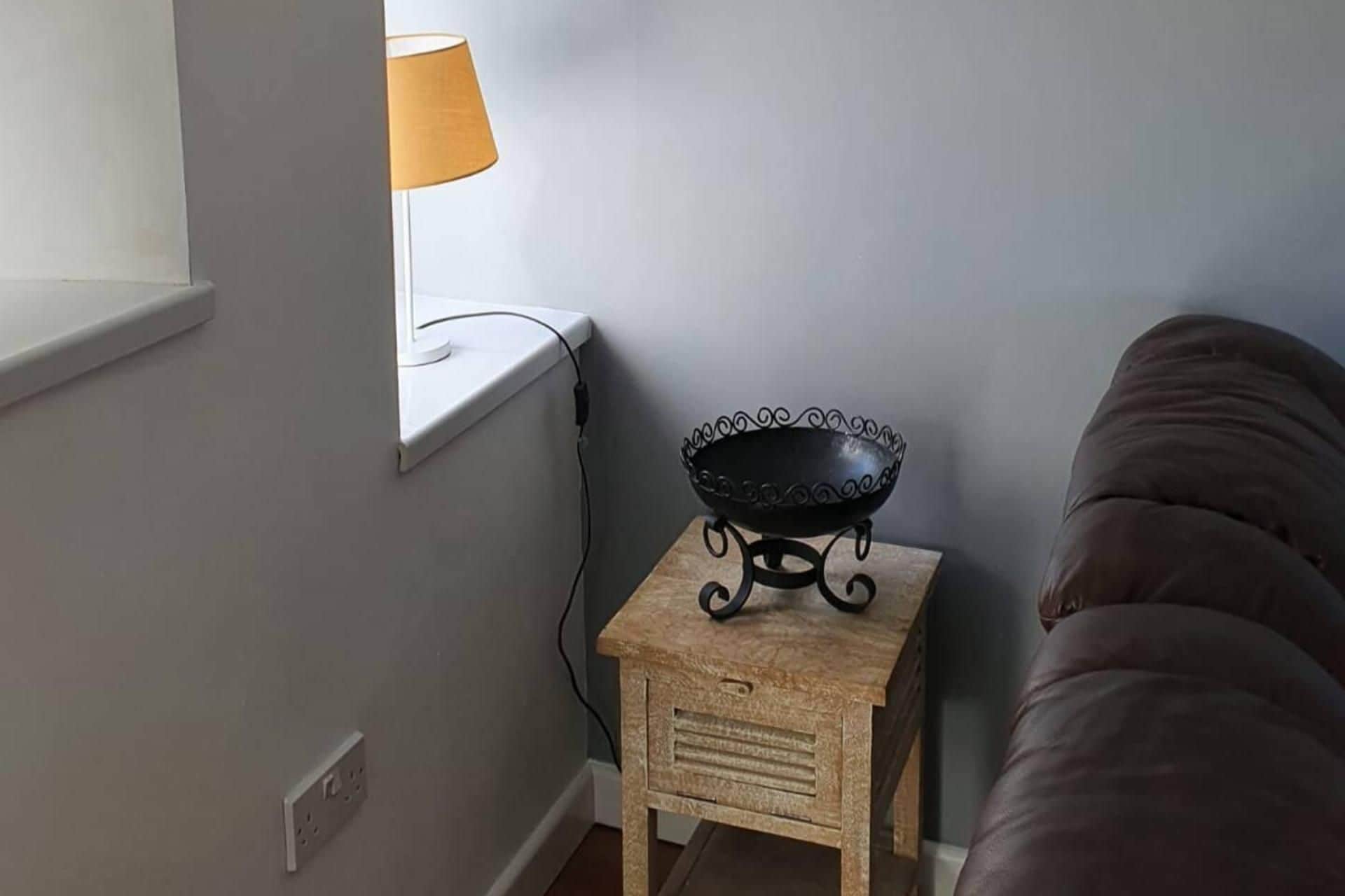 Un taburete de madera pequeño con un bol metálico encima, una lámpara en el alféizar de una ventana y un sofá marrón.
