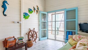 A room with a wooden floor, a bed with floral bedding, a wooden chair, a small table with books and a basket, a wall-mounted TV, and a door with a blue frame. - Casa Beira Mar enseada dos Corais-SH098D (Cabo de Santo Agostinho)