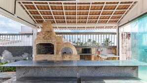 An outdoor kitchen with a brick oven, stone countertops, and wooden benches. - Casa Beira Mar enseada dos Corais-SH098D (Cabo de Santo Agostinho)