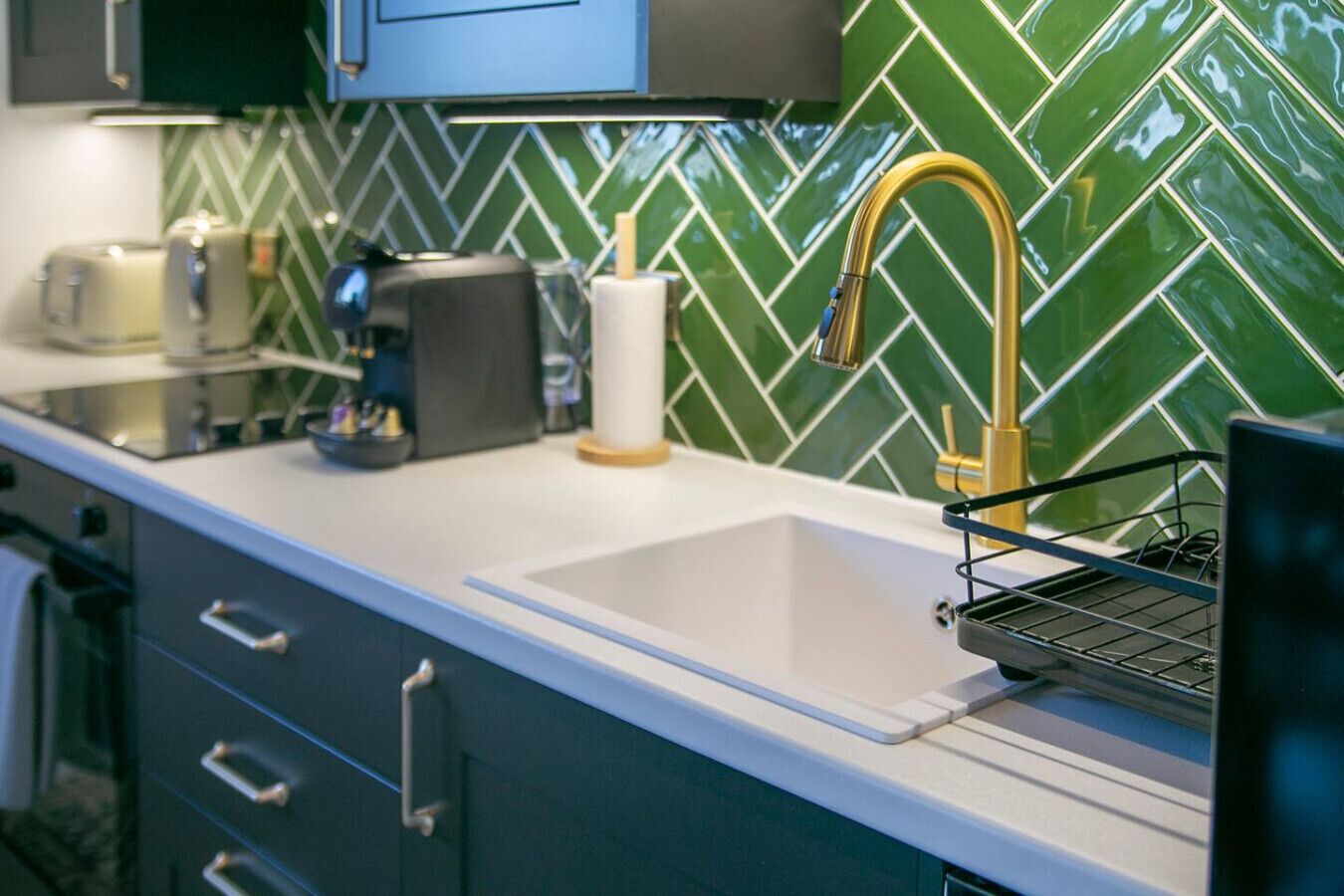 A kitchen with a white countertop, a gold faucet, and a green tiled backsplash.