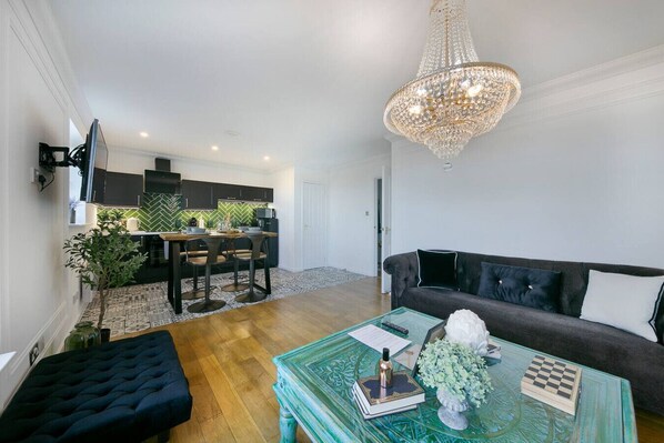 A modern living room with a dark sofa, a glass coffee table with books and a chess set, a chandelier, and a kitchen in the background.