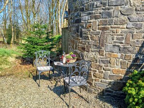 A stone wall with two metal chairs and a table, surrounded by gravel and greenery.