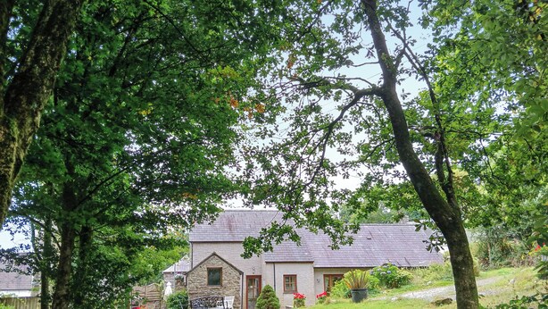 Une maison en pierre avec un toit en pente et un petit porche à l’avant, entourée d’une végétation luxuriante et d’une cour gazonnée.